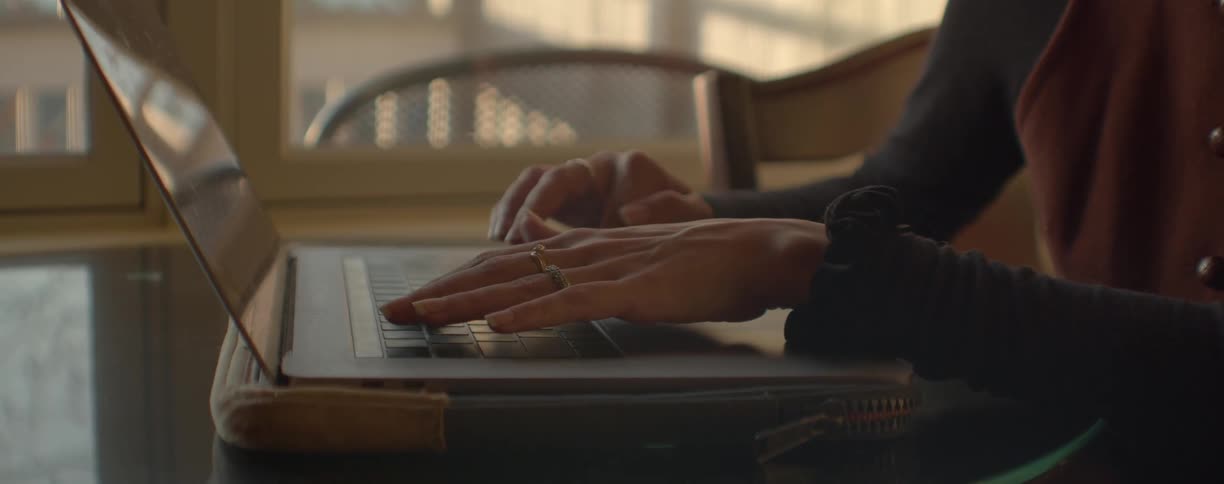 Woman with black shirt and brown vest works on laptop next to windows in room