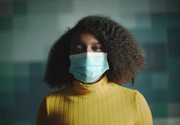 A young woman wearing a surgical mask appears to smile as she stands in a room