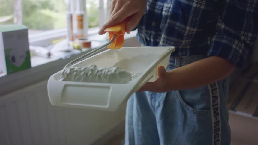 A person rolls a paint roller in white paint in a tray to prepare it for use