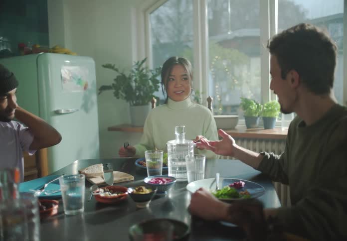 A group of people talk with one another while eating together at a dining table in a home kitchen during the day