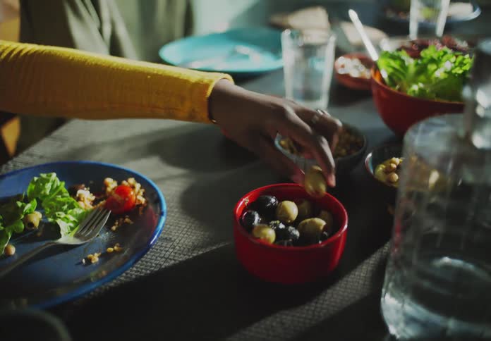Woman with yellow shirt sitting at dining table next to man with goatee and green sweater takes olive from red bowl in the kitchen