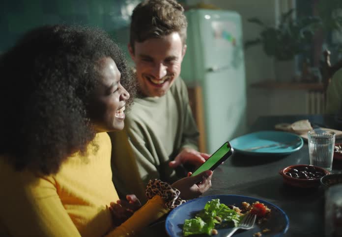 A woman sits at a table next to a man and laughs as she holds her cell phone and they talk