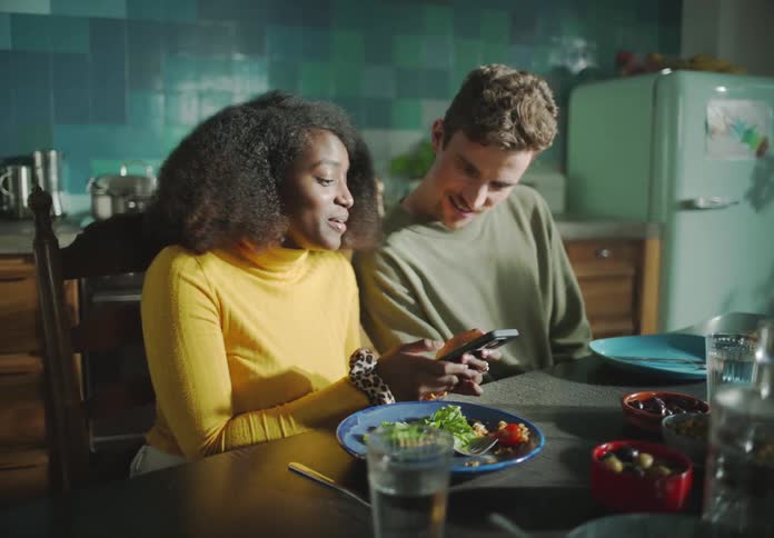 A man and woman look at the womans cell phone and laugh while sitting at a dining table with other people