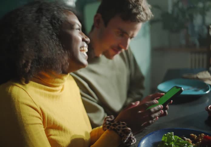 A young woman shows the screen of her smartphone to a man sitting next to her at a dining table and they laugh together