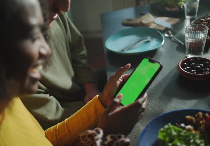 A man and woman talk and laugh as they look at a cell phone while seated at a table of food