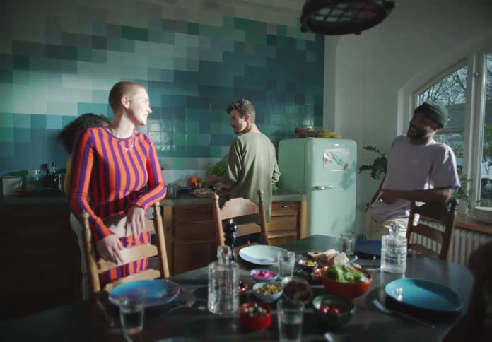 A group of young men and woman talk together in a kitchen while one man preps food