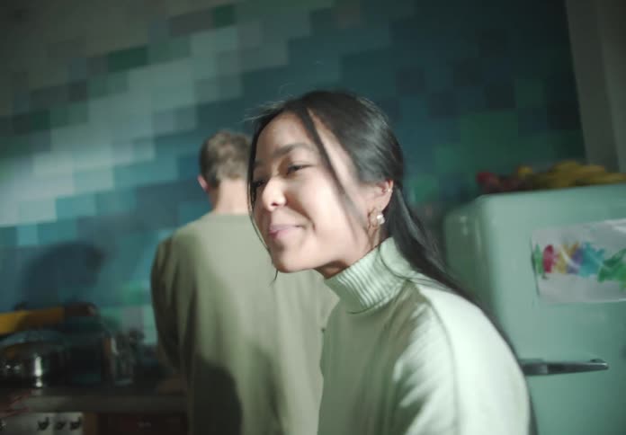 Woman with straight hair white turtleneck sweater talks to friends while man with green shirt and woman with frizzy hair and yellow shirt are cooking in the kitchen at home