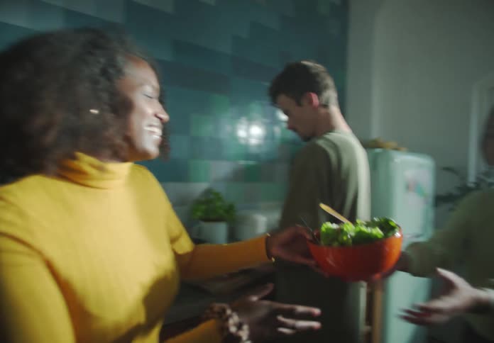 A woman passes a salad bowl to another woman in a kitchen