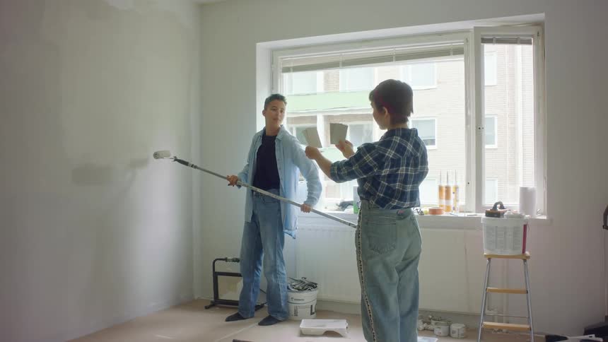 A woman holds up several paint color swatches and a man nods his approval before continue to roll paint on a wall