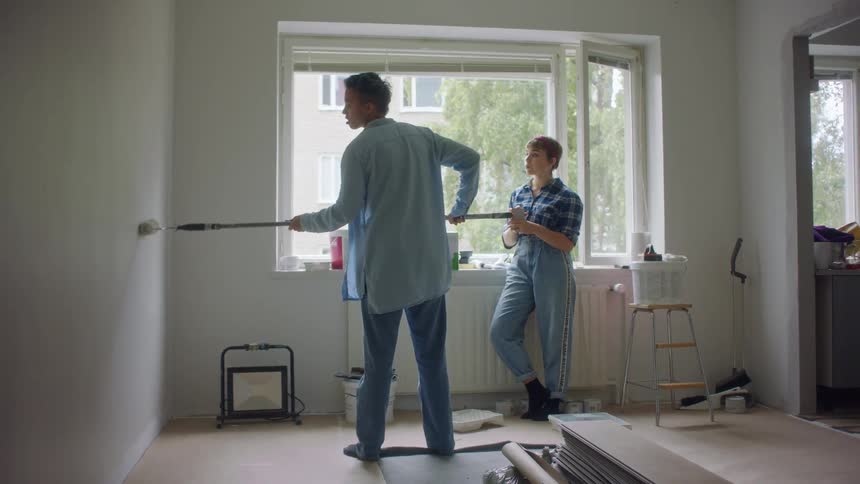 A person uses a paint roller to paint the wall of a home and a woman watches