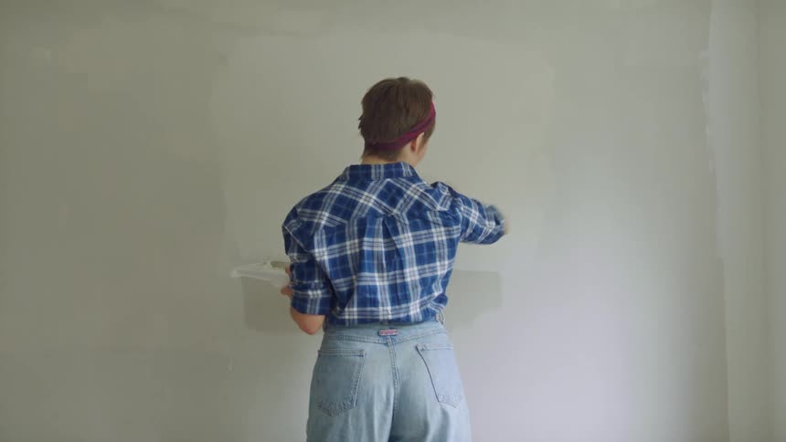 Woman with short brown hair blue plaid shirt and jeans paints wall white in room