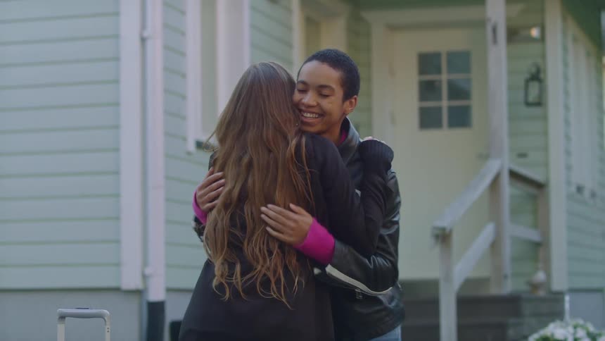 A woman runs down the steps of a house to greet another woman with a hug and smile