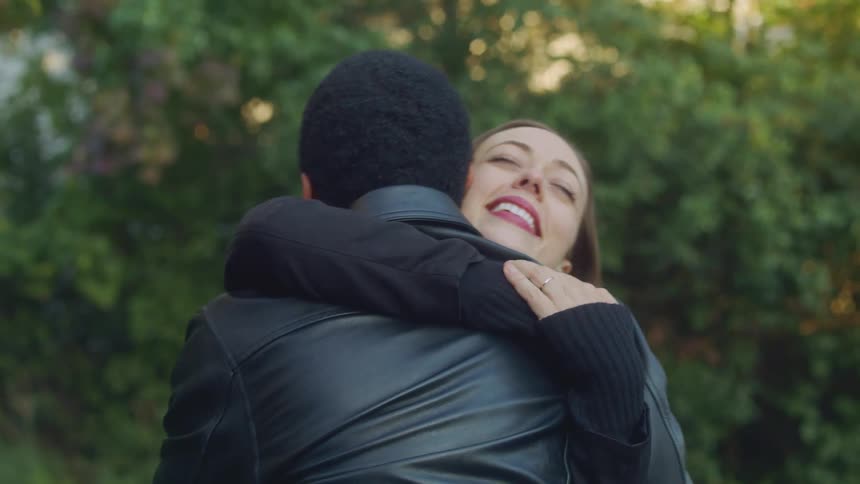 A woman drops the handle of her suitcase to hug her friend tightly as they greet each other in the driveway