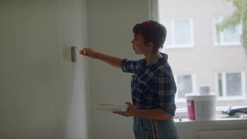 A woman uses a small paint roller to paint a spot on a wall