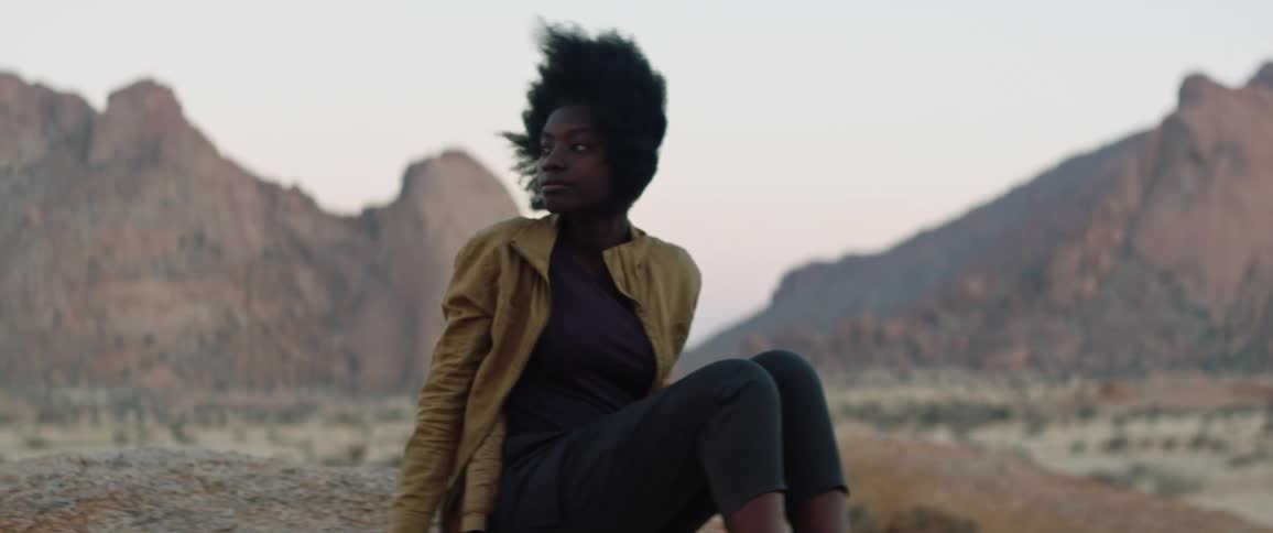 Wind blows womans hair as she sits with scrub field and mountains in the background