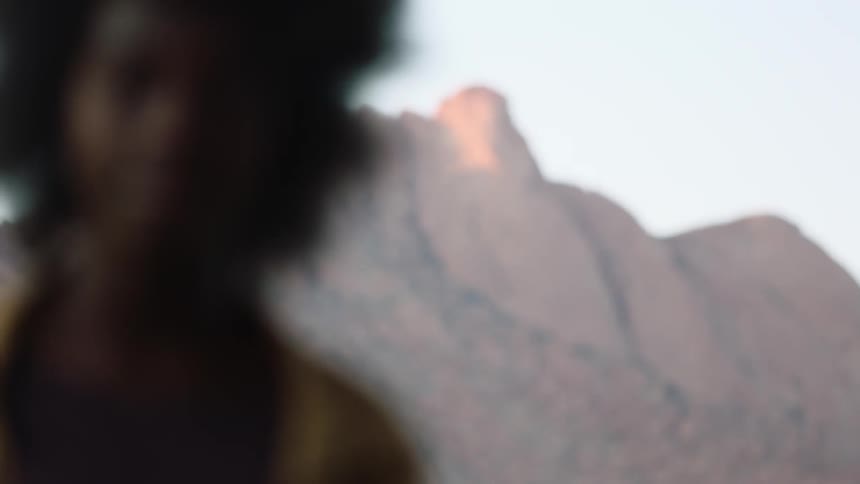 Young black woman with afro standing in the desert with mountains in background looks into the camera