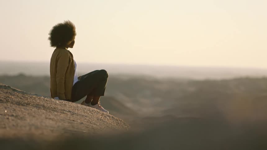 Woman sits on ground overlooking valley under a red tinted sky