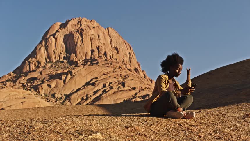 Black female sitting in front of large rock formation face timing on cell phone