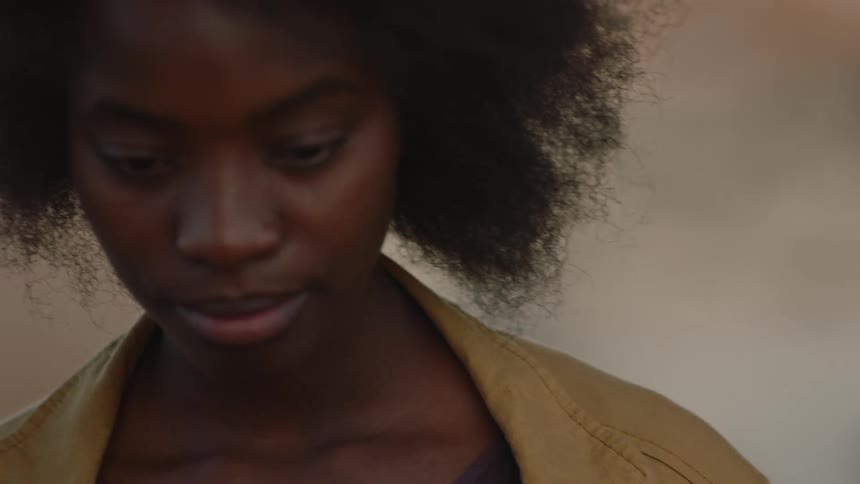 Young black woman walks outside with her hands in her coat pocket and then looks up at the camera