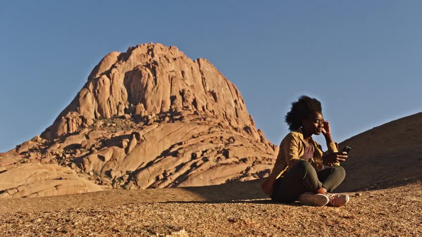 A young black woman is sitting on the ground in front of a mountain talking on her cell phone