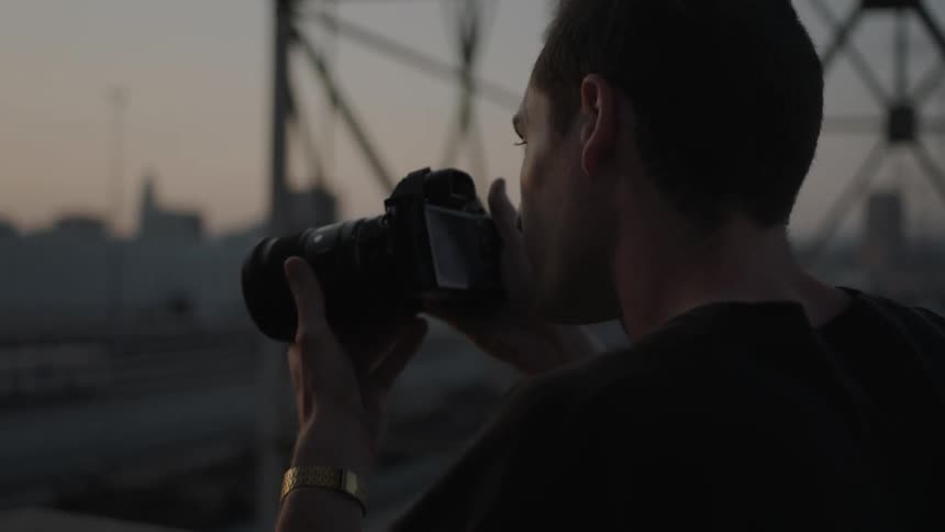 A man slowly lifts a camera to take a photo of a city at sunset