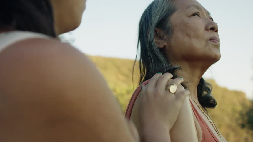 Woman with white sleeveless shirt rubs womans shoulder wearing sleevless red shirt looking up outdoors