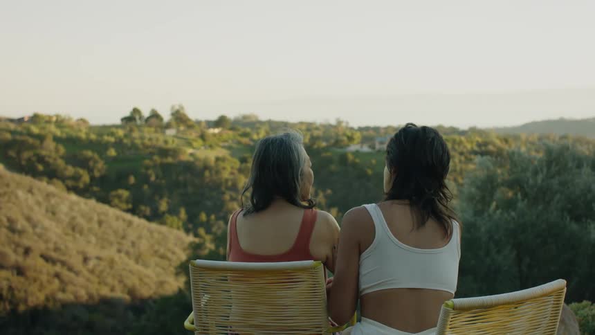 Woman in white swimsuit puts on sun cream on womans shoulder in red swimsuit sitting in armchair overlooking hills