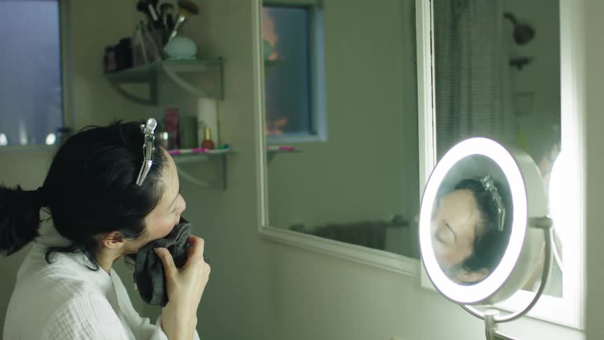 Woman sitting in front of a mirror cleans the skin on her face with a damp wet cloth