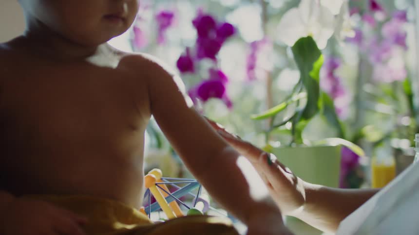 A woman at home rubs her infant sons arms as another person reads a book