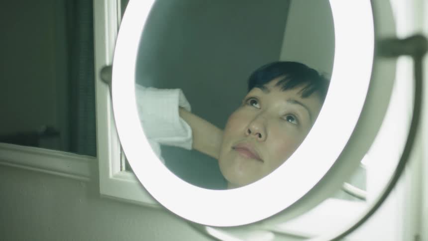 An asian woman is looking in a round mirror with a light around it as she fixes her hair