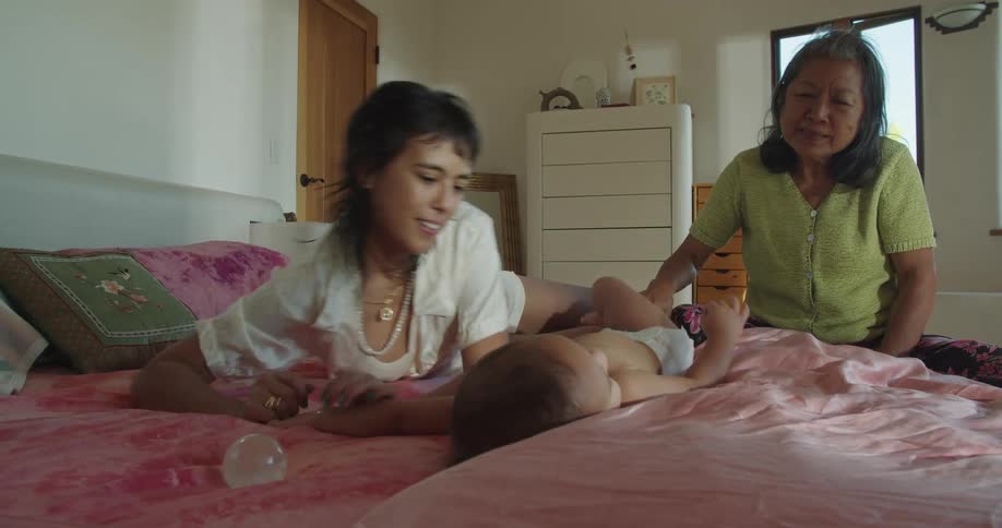 A woman lying on her bed playing with her baby while grandma smiles and caresses his leg