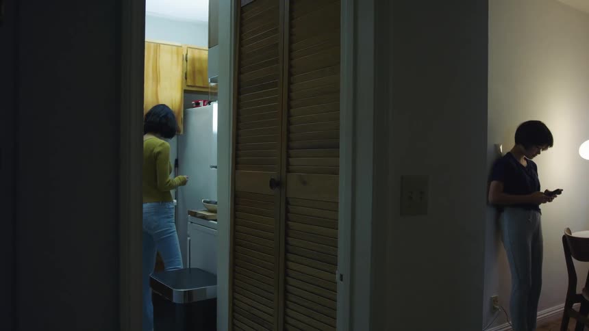 A woman prepares food in a kitchen while her daughter stands in the next room and texts on her cell phone