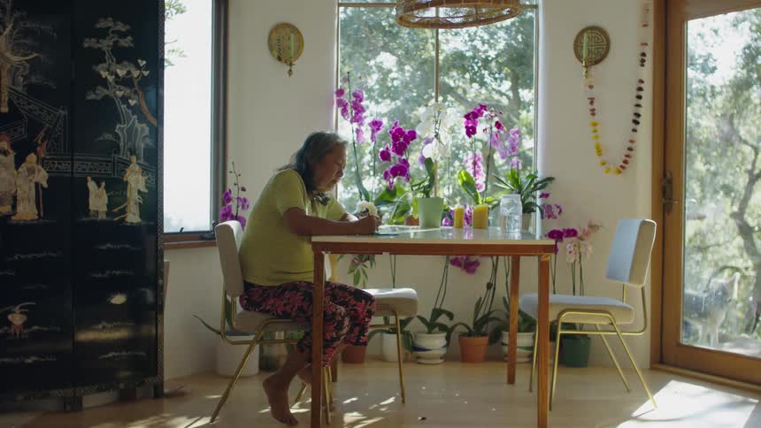 An elderly woman sits at a table in a home