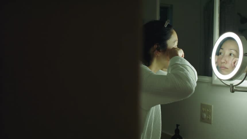 Woman stands in front of makeup mirror in the bathroom massaging lotion into her face