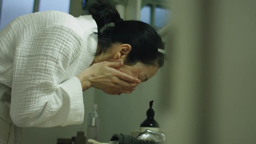A woman bent over the bathroom sink washes her face and rinses with water