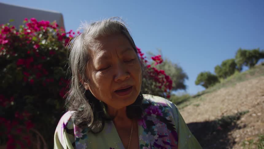 An elderly woman singing outside near a flowering bush