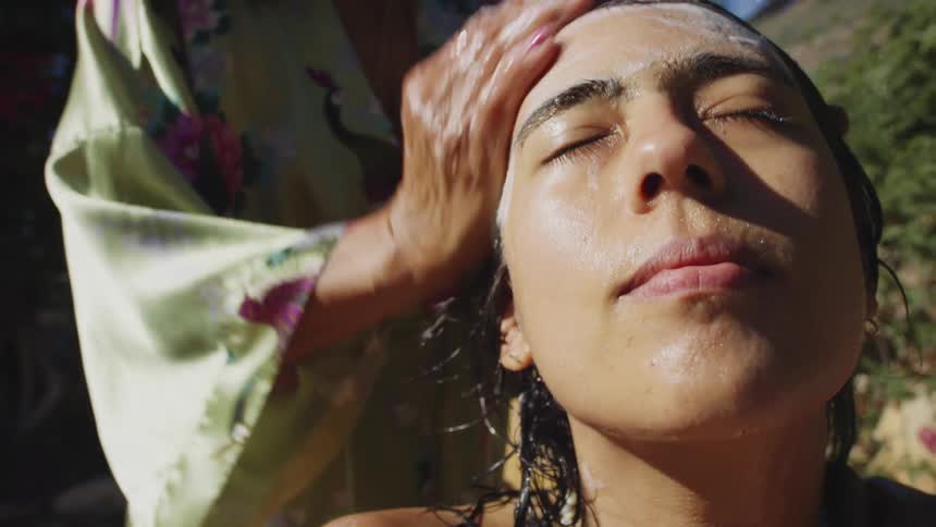 An elderly woman wearing a bathrobe washes her daughters hair outside in the bright sunshine