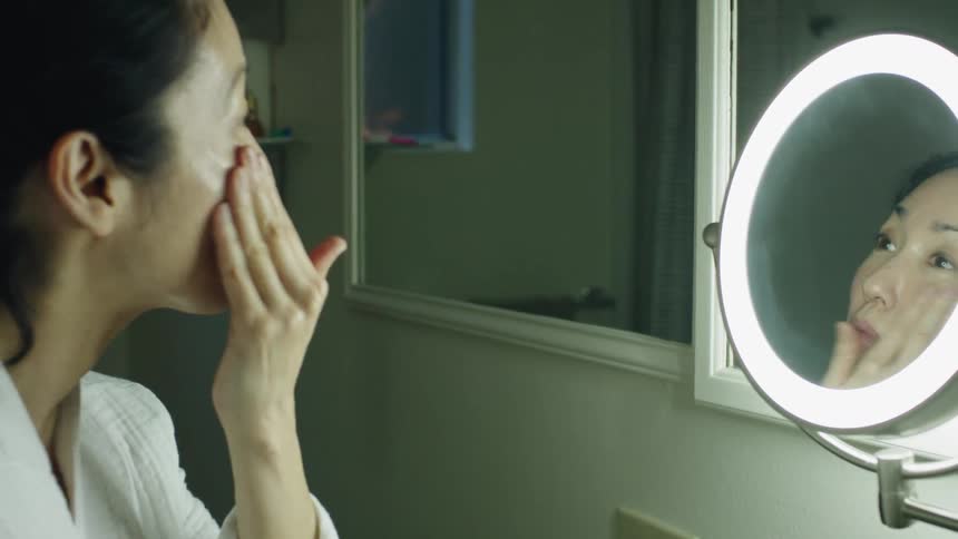 Woman with white bathrobe puts cream on her face in bathroom