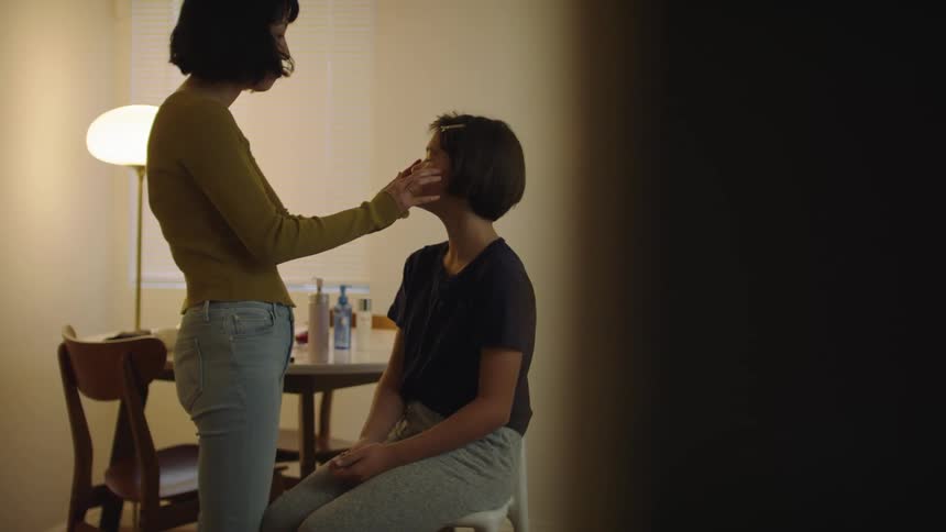Woman applies creams or makeup on the face of a friend sitting on a chair inside a room