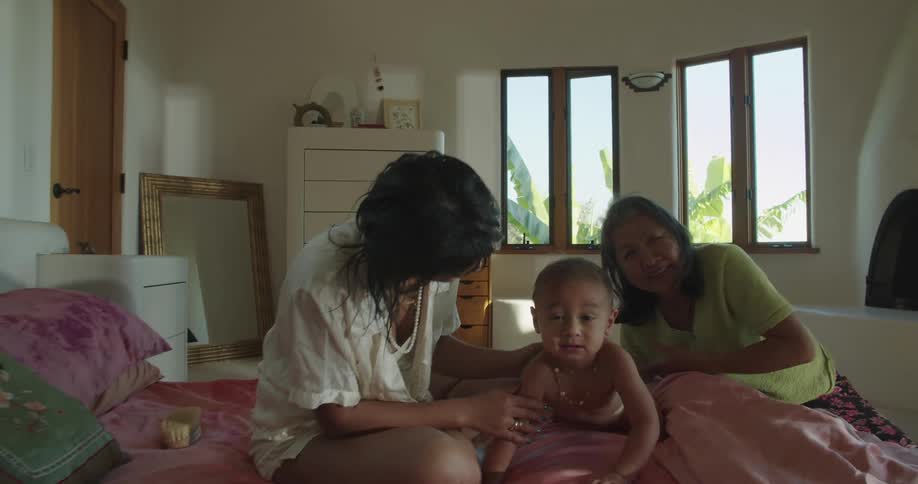 A woman sits on a bed with her mother and baby son as she rubs his back and they all laugh