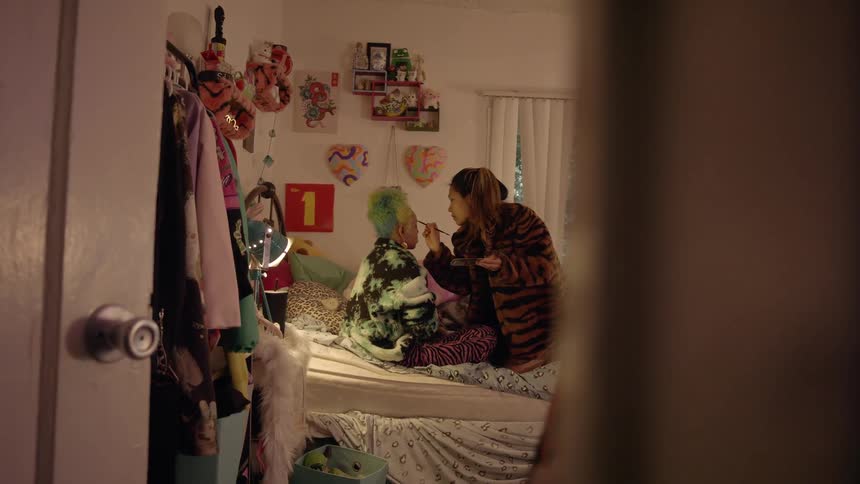 Two teenagers sit on a bed as one puts make up on the other