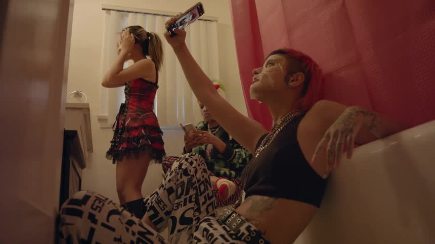 A teenage girl with a face mask on leans against a bathtub and takes a selfie on her phone while another girl sits and texts and another girl applies a face mask