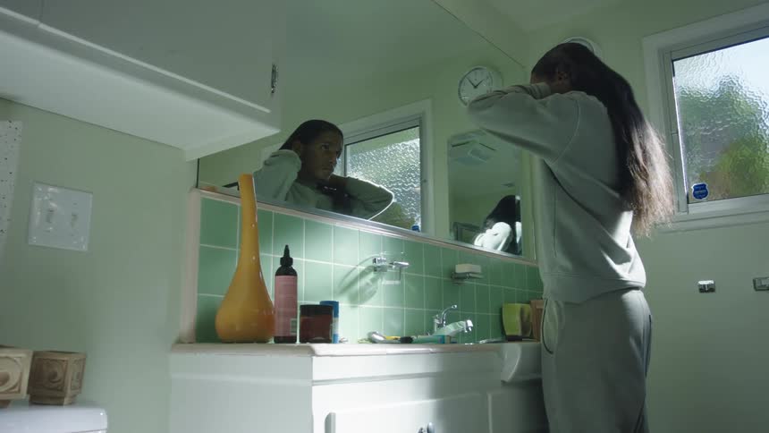 A teenage stands in front of a mirror in a bathroom and puts a necklace on
