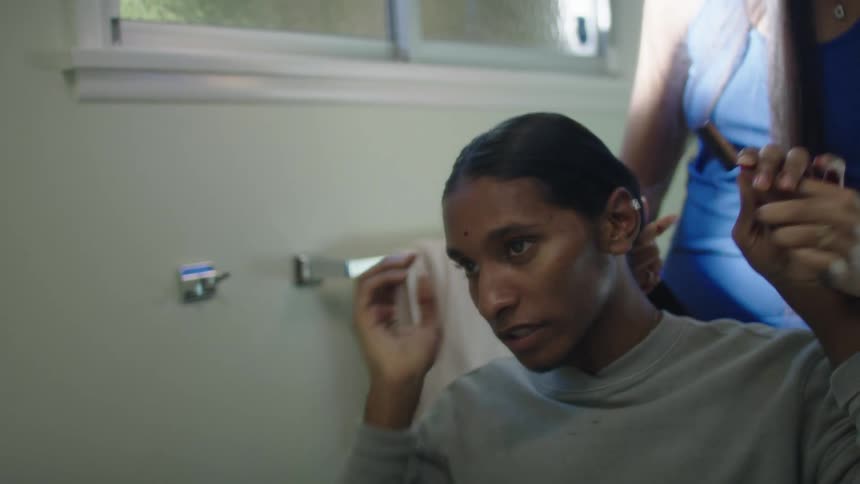 Woman with long straight hair and blue dress combs mans long hair in bathroom
