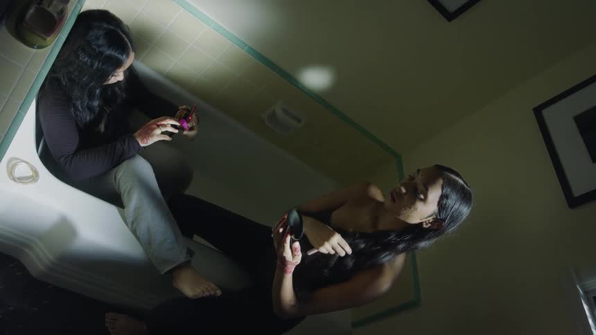 A teenage girls sits fully clothed in a bathtub while a woman sits on the edge and moves a makeup compact around