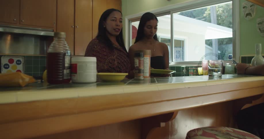 Mother and daughter work in kitchen as other women sit on stools at counter