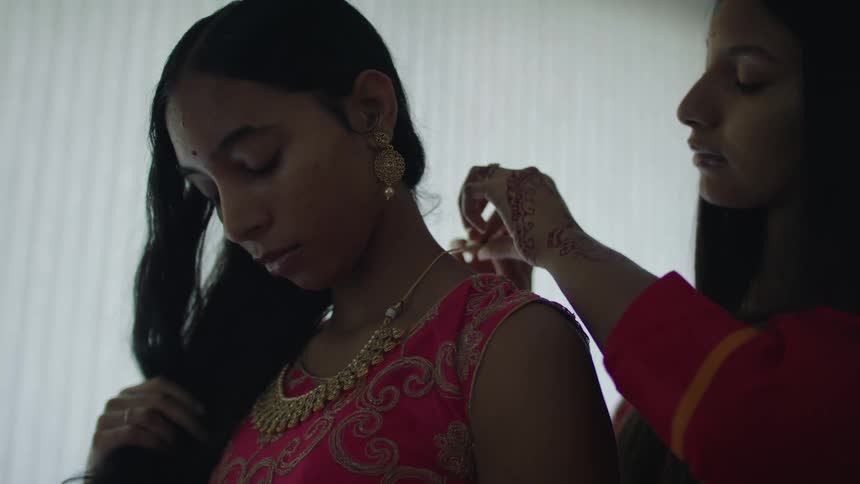 Two young women are standing while one fastens a necklace on the other