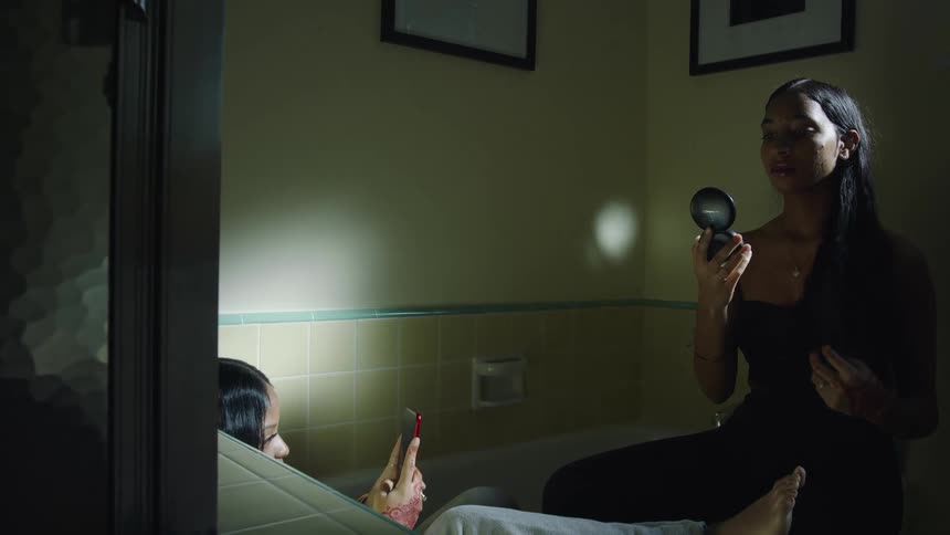 Friends sitting on an empty bathtub applying makeup and taking selfies