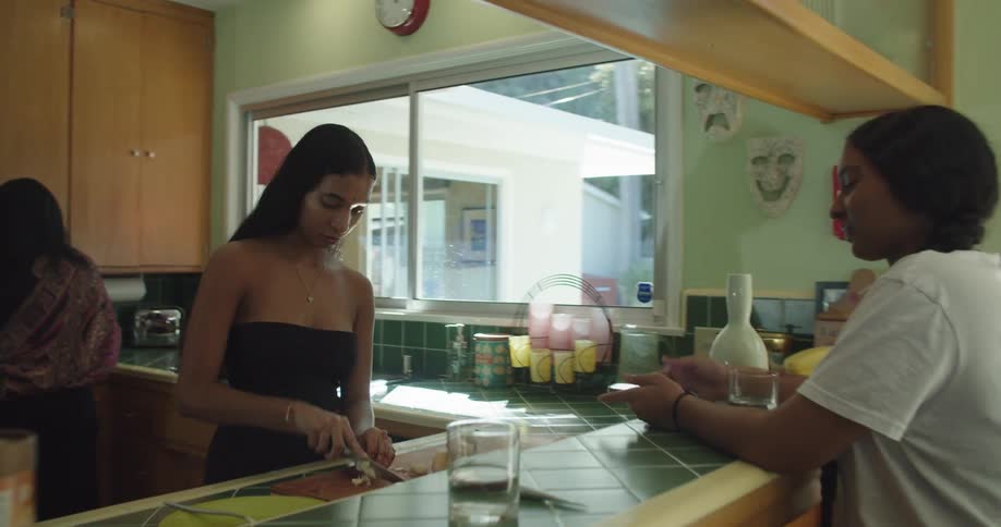 Woman with straight hair black dress cuts vegetable on cutting board and woman with white t shirt drinks a glass of water in the kitchen