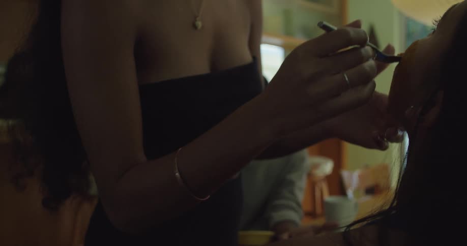 Woman helps another put on makeup while another woman stands in kitchen